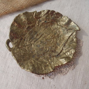 May include: A gold-colored metal leaf-shaped dish with a textured surface. The dish has a small handle on the side.