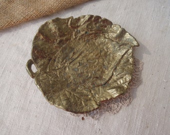 May include: A gold-colored metal leaf-shaped dish with a textured surface. The dish has a small handle on the side.
