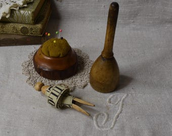 May include: A wooden pincushion with a brown fabric top and colorful pins, a wooden thimble, and a vintage measuring tape on a white linen surface.