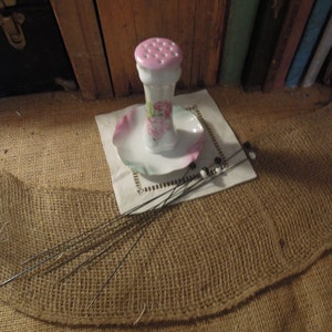 May include: A vintage porcelain pincushion with a floral design and a pink top. The pincushion is sitting on a white saucer with a ruffled edge. There are several black and white pins on a white doily next to the pincushion.