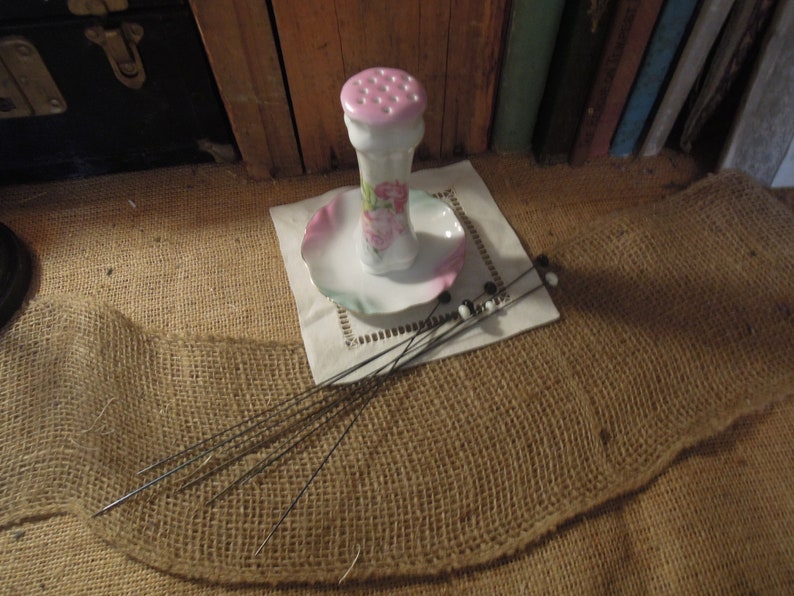 May include: A vintage porcelain pincushion with a floral design and a pink top. The pincushion is sitting on a white saucer with a ruffled edge. There are several black and white pins on a white doily next to the pincushion.