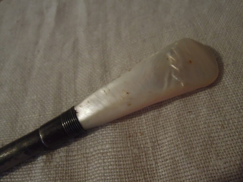 May include: A white mother-of-pearl handle with a silver metal shaft. The handle has a decorative pattern.