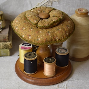 May include: A vintage sewing kit featuring a floral pin cushion, wooden base, and spools of thread in various colours. The pin cushion is light brown with a floral pattern.