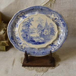 May include: A vintage blue and white porcelain plate with a landscape scene. The plate has a scalloped edge and is displayed on a small wooden stand. The design features a church, trees, and figures.