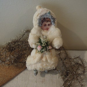 May include: A vintage white cotton doll ornament with a painted face, wearing a white fur coat and holding a bouquet of pink flowers.