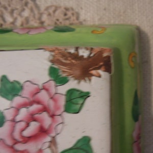 Op de afbeelding: Een groene en witte keramische tegel met een bloemenmotief. De tegel heeft een roze bloem met groene bladeren en een bruine vogel op de achtergrond. De tegel heeft een gebroken rand.