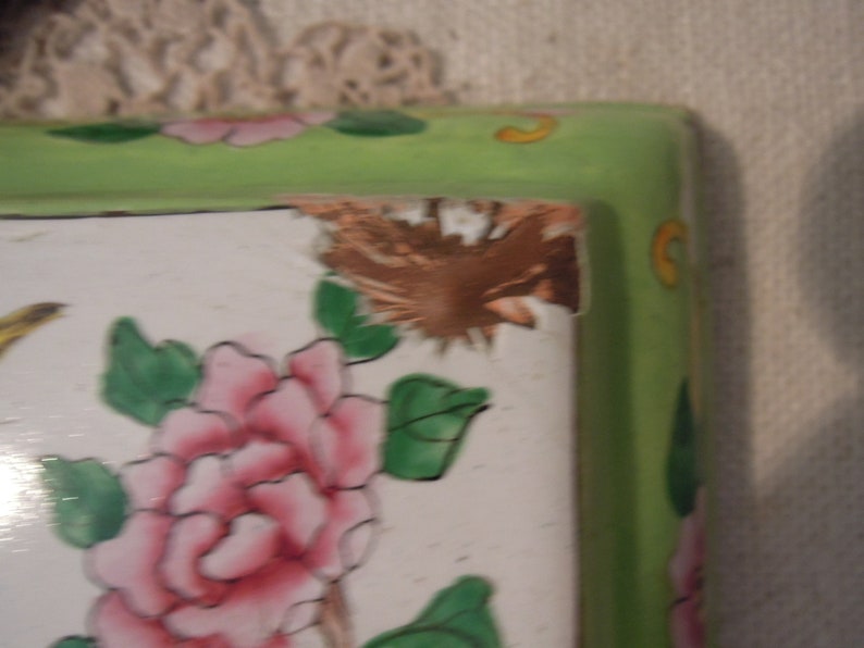 Op de afbeelding: Een groene en witte keramische tegel met een bloemenmotief. De tegel heeft een roze bloem met groene bladeren en een bruine vogel op de achtergrond. De tegel heeft een gebroken rand.