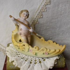 May include: A porcelain figurine of a cherub rowing a yellow and white boat with floral details. The cherub is wearing a pink and white dress and holding a wooden oar.