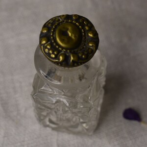 May include: A clear glass bottle with a decorative, ornate, gold-colored metal lid. The bottle has a faceted design.