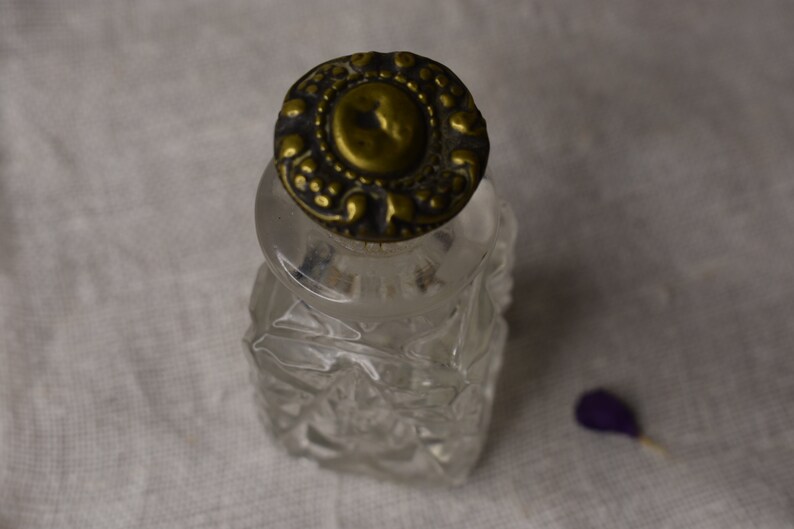 May include: A clear glass bottle with a decorative, ornate, gold-colored metal lid. The bottle has a faceted design.