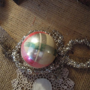 May include: A vintage Christmas ornament with a silver, green, and pink geometric design. The ornament is surrounded by silver tinsel.