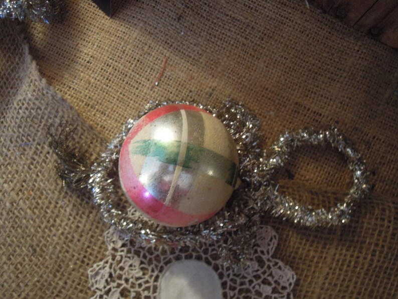 May include: A vintage Christmas ornament with a silver, green, and pink geometric design. The ornament is surrounded by silver tinsel.