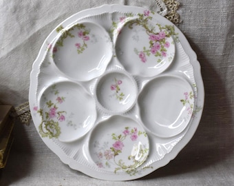 May include: White porcelain oyster plate with six individual compartments and a central well. Each compartment and the well are decorated with pink rose floral patterns and green accents. The plate has a scalloped edge.