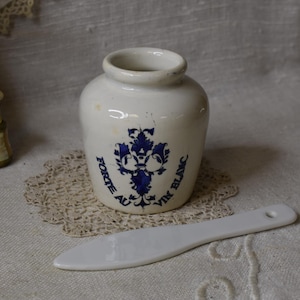 May include: A small, off-white ceramic jar with a blue floral design and text. A white, spatula-like tool rests in front of the jar. The jar sits on a lace doily, with other vintage items in the background.