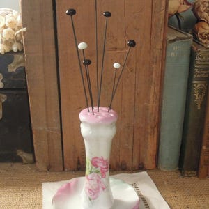 May include: A white porcelain pincushion with a pink floral design and a matching saucer. The pincushion has a tall, slender shape and is decorated with pink roses. The pincushion is filled with black and white pins.