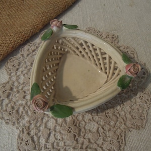 Puede incluir: Una peque&ntilde;a cesta de cer&aacute;mica blanca con un dise&ntilde;o tejido y detalles de rosas rosas. La cesta tiene forma triangular y tiene una est&eacute;tica delicada y femenina.