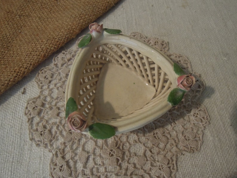 Puede incluir: Una peque&ntilde;a cesta de cer&aacute;mica blanca con un dise&ntilde;o tejido y detalles de rosas rosas. La cesta tiene forma triangular y tiene una est&eacute;tica delicada y femenina.