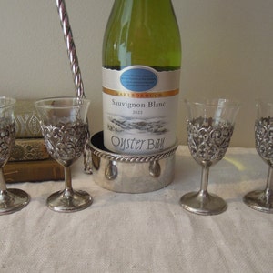 Könnte beinhalten: Eine Flasche Marlborough Sauvignon Blanc 2023 steht in einem silbernen Weinkühler. Vier kleine, klare Weingläser mit verzierten silbernen Filigranbasen sind davor angeordnet. Im Hintergrund befindet sich ein silberner Flaschenöffner.