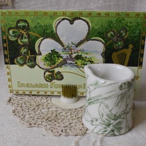 Puede incluir: Una postal vintage con un diseño de trébol y las palabras "Ireland Forever!" se muestra detrás de una pequeña jarra blanca con patrones florales verdes. Un soporte floral dorado y un tapete de encaje están al frente.