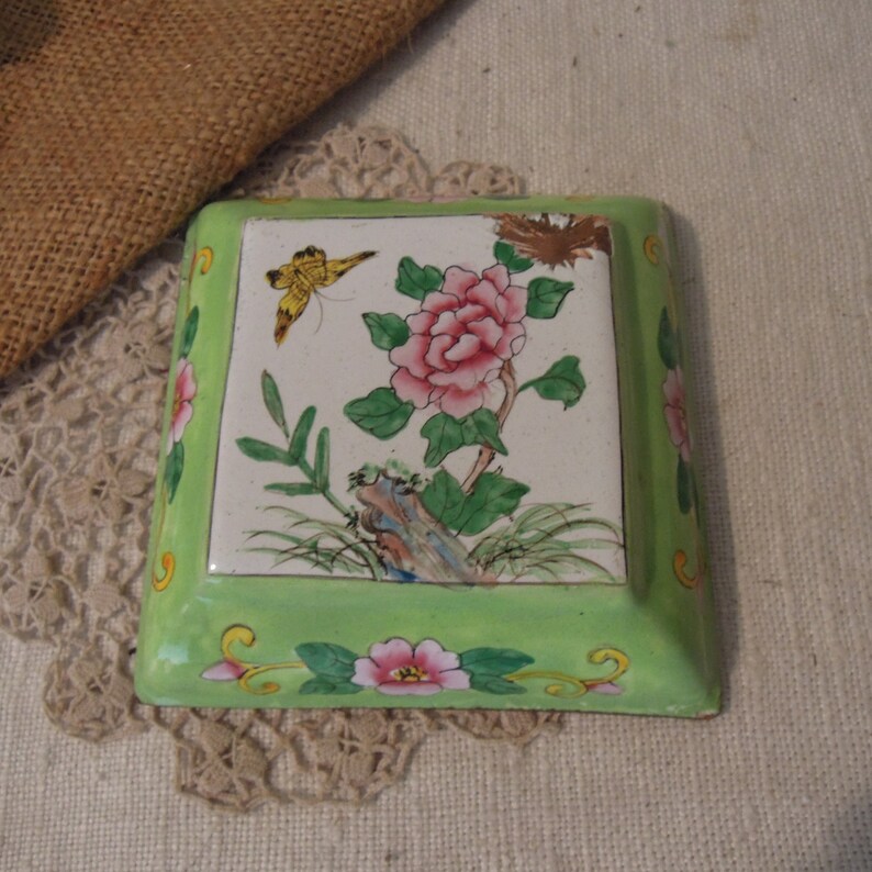 Op de afbeelding: Een vierkante groene en witte keramische sieradenbox met een bloemenmotief. De doos heeft een roze bloem met groene bladeren en een gele vlinder.