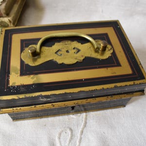 May include: An antique black and gold metal box with a brass handle and decorative accents. The box has a rectangular shape with a hinged lid and a keyhole. The box is likely used for storing small valuables.