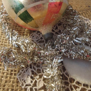 May include: A vintage glass Christmas ornament with a gold, red, green, and white geometric pattern. The ornament is surrounded by silver tinsel and a white crocheted doily.