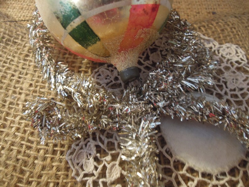 May include: A vintage glass Christmas ornament with a gold, red, green, and white geometric pattern. The ornament is surrounded by silver tinsel and a white crocheted doily.