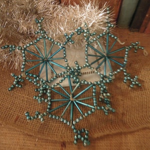 May include: A set of three teal blue beaded snowflake ornaments. The snowflakes are made of wire and beads and are strung together with a loop for hanging.