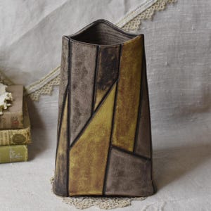 Puede incluir: Un jarrón alto y rectangular con un diseño geométrico. El jarrón presenta un mosaico de formas marrones y amarillas delineadas en negro. El jarrón tiene una parte superior ligeramente ensanchada y una superficie texturizada, ideal para la decoración del hogar.