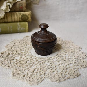 May include: A small, dark brown wooden container with a lid, resting on a cream-colored, lace doily. The container has carved details around the base. The background includes antique books and fabric.