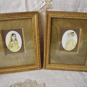 May include: Two rectangular gold-toned frames with beaded edges, each containing an oval-shaped portrait painting. One portrait depicts a person in a yellow garment and head covering, while the other shows a person wearing a yellow turban and attire.