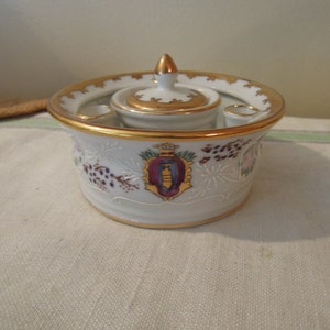 May include: A white porcelain inkwell with a gold trim and a floral design. The inkwell has a small lidded jar and two small inkwells.