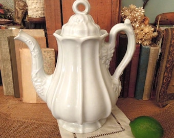 May include: A white ceramic teapot with a fluted body, a matching lid featuring a ring-shaped handle, and a curved spout. The teapot is decorated with embossed grape and vine motifs on the spout and handle, and it sits on a square cloth.