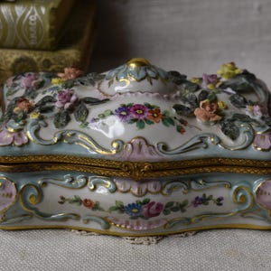 May include: A white porcelain trinket box with a hinged lid. The box is decorated with a floral design in shades of pink, blue, and green. The lid features a raised floral design and a gold trim.