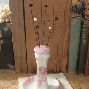 May include: A white porcelain pincushion with a pink floral design and black and white glass heads on the pins. The pincushion is sitting on a white doily.
