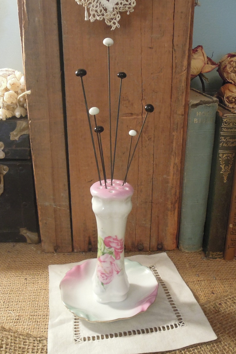 May include: A white porcelain pincushion with a pink floral design and black and white glass heads on the pins. The pincushion is sitting on a white doily.