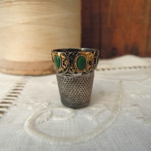 May include: A vintage silver thimble with a gold-colored band featuring green oval-shaped embellishments. The thimble has a textured, perforated surface and sits on a white, patterned cloth. A spool of thread is in the background.