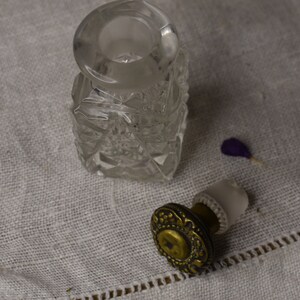 May include: A clear glass bottle with a textured surface and a gold metal stopper. The bottle is sitting on a white fabric surface.