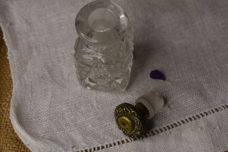 May include: A clear glass bottle with a textured surface and a gold metal stopper. The bottle is sitting on a white fabric surface.
