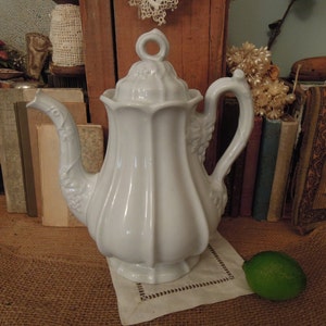 May include: White porcelain teapot with a fluted body and a floral design. The teapot has a lid with a knob and a spout. The teapot is sitting on a burlap surface.