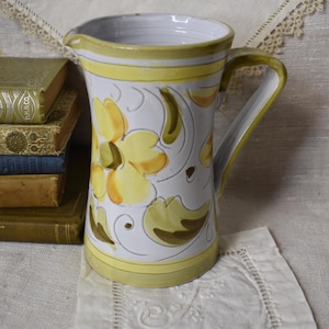 以下が含まれることがあります： 黄色と緑の花柄の白い陶器のピッチャー。ピッチャーには取っ手と注ぎ口があります。