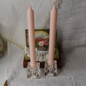 Può includere: Due candele affusolate rosa pallido in portacandele quadrati di cristallo trasparente. Sullo sfondo una stampa floreale vintage e un centrino in pizzo. La scena è ambientata su una superficie bianca e strutturata.