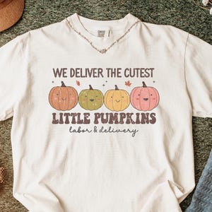 May include: Cream-colored t-shirt with the text "WE DELIVER THE CUTEST LITTLE PUMPKINS labor & delivery" and four smiling pumpkin illustrations in orange, green, yellow, and pink. The shirt is laid on a textured surface with accessories.