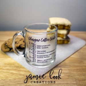 May include: A clear glass mug with a black printed Glasgow Coffee Scale. The scale measures from 3 to 15 and includes descriptions of different levels of consciousness. The mug is sitting on a white cutting board with a stack of cookies and a stack of pastries in the background.