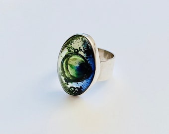 May include: A silver ring with an oval-shaped green, blue, and white resin inlay. The resin has a swirling pattern and small air bubbles.
