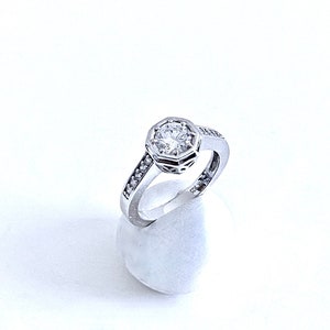 May include: A silver ring with a large, clear, round gemstone surrounded by smaller gemstones. The ring is on a white display stand.