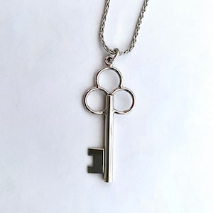May include: A silver-colored key pendant necklace. The key has a classic design with a long stem and a three-circle top. The chain is a delicate, woven style. The necklace is displayed against a white background.
