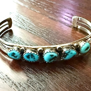 May include: A silver cuff bracelet featuring five oval-shaped turquoise stones with dark matrix patterns. The metal band has a polished finish with decorative twisted wire detailing along the sides and small silver beads between each stone setting.