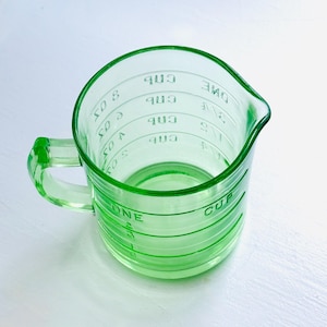 May include: A vintage green glass measuring cup with a handle and a pouring spout. The cup has measurement markings in cups and fractions of a cup. The cup is made of a translucent green glass.