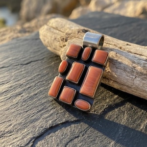 May include: A silver pendant with a rectangular black base, featuring multiple rectangular and oval-shaped orange-red stones. The pendant has a silver loop at the top for a chain. The background includes a piece of driftwood and a dark, textured surface.
